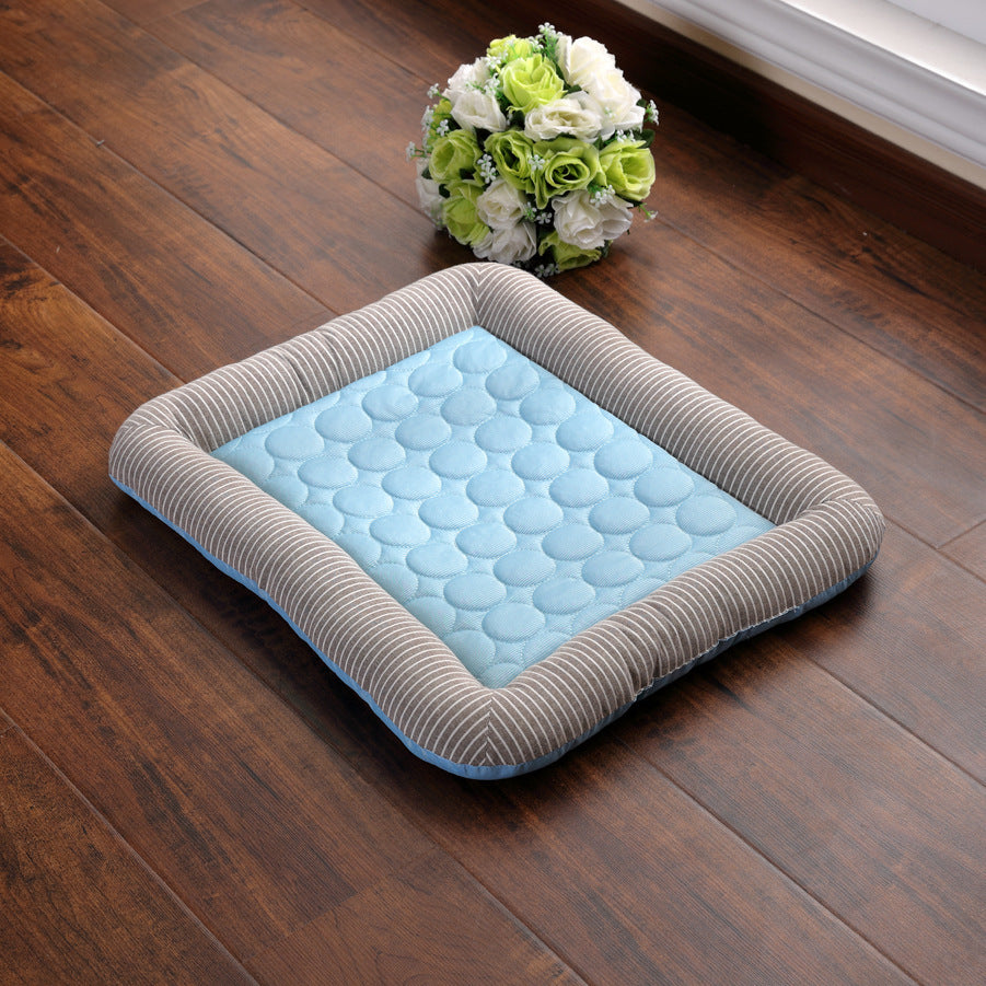 Blue quilted cooling pet bed mat with striped gray cushioned edges placed on wooden floor next to bouquet of white and green flowers.