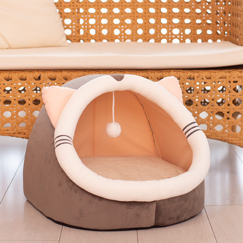 Gray and peach plush cat cave bed with cat ears and hanging pompom under wicker chair