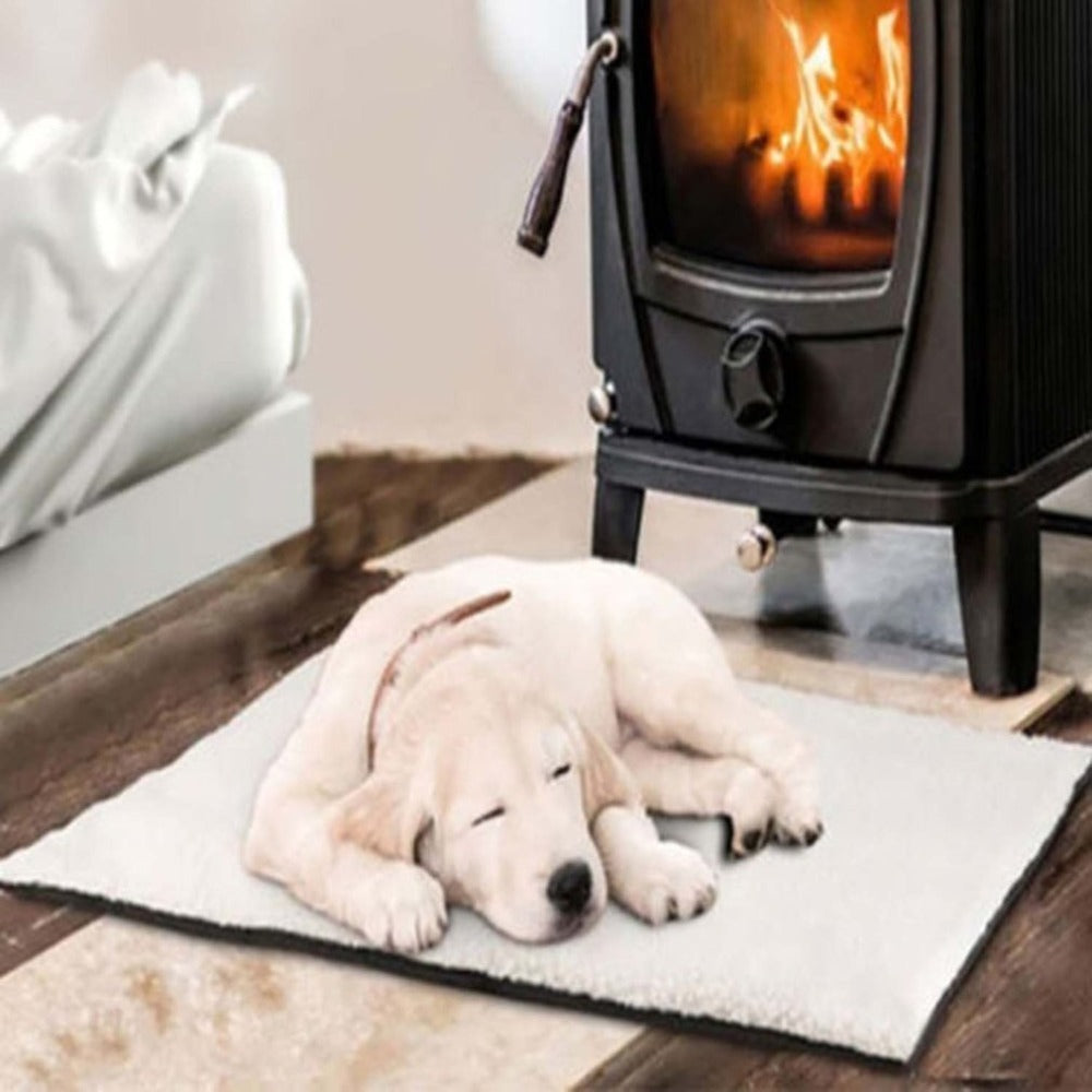Light-colored puppy lying on a soft white self-heating pet blanket mat near a black fireplace with fire burning inside.