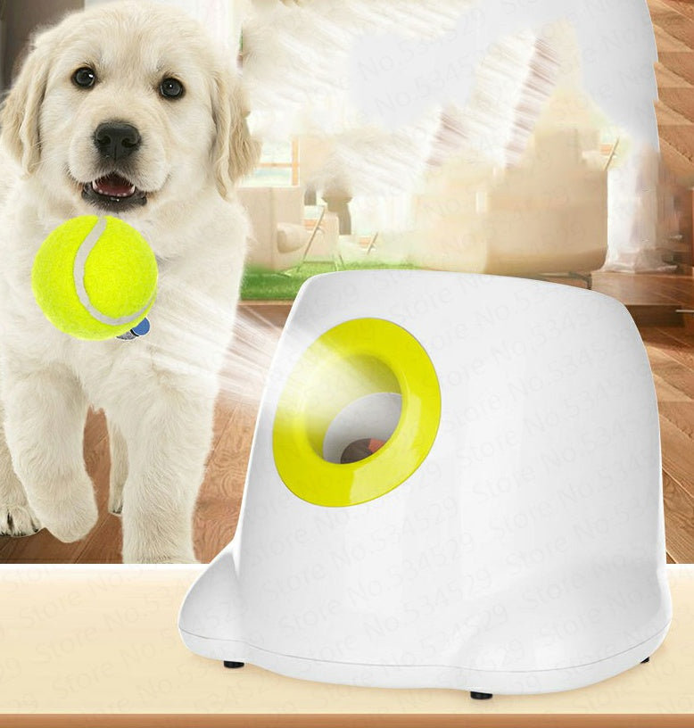 White automatic ball launcher with yellow-green launching opening beside happy golden retriever catching a tennis ball indoors