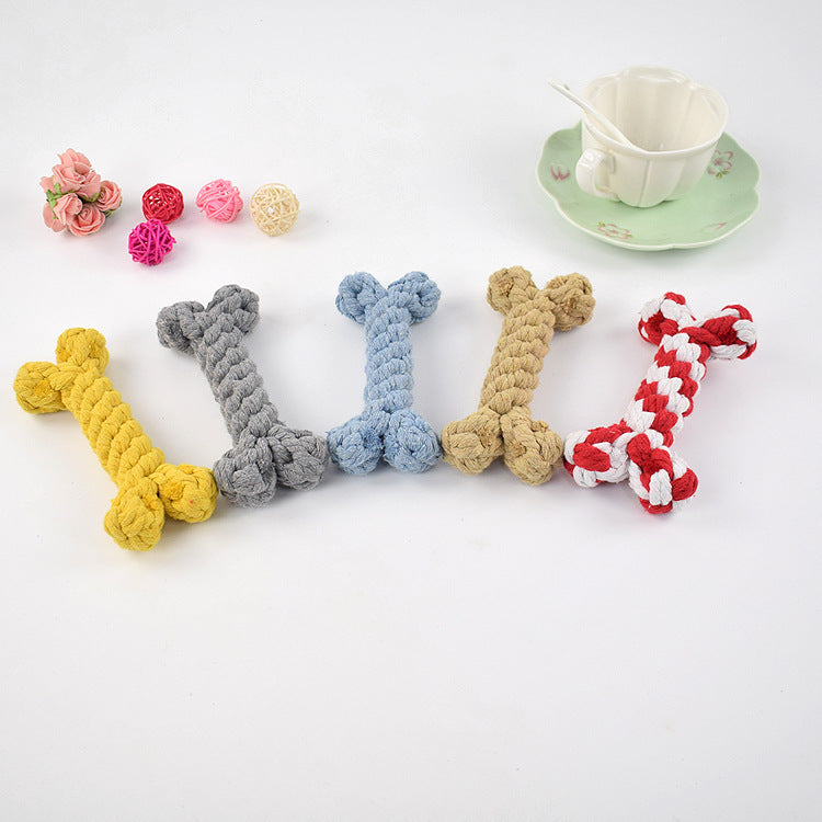 Five bone-shaped braided cotton rope dog toys in yellow, gray, blue, tan, and red with white, lined up on white surface with decorative balls and teacup
