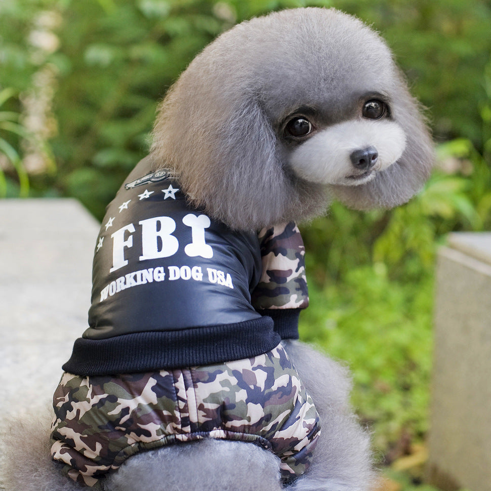Gray toy poodle wearing camo FBI jumpsuit looking back, outdoors with greenery background
