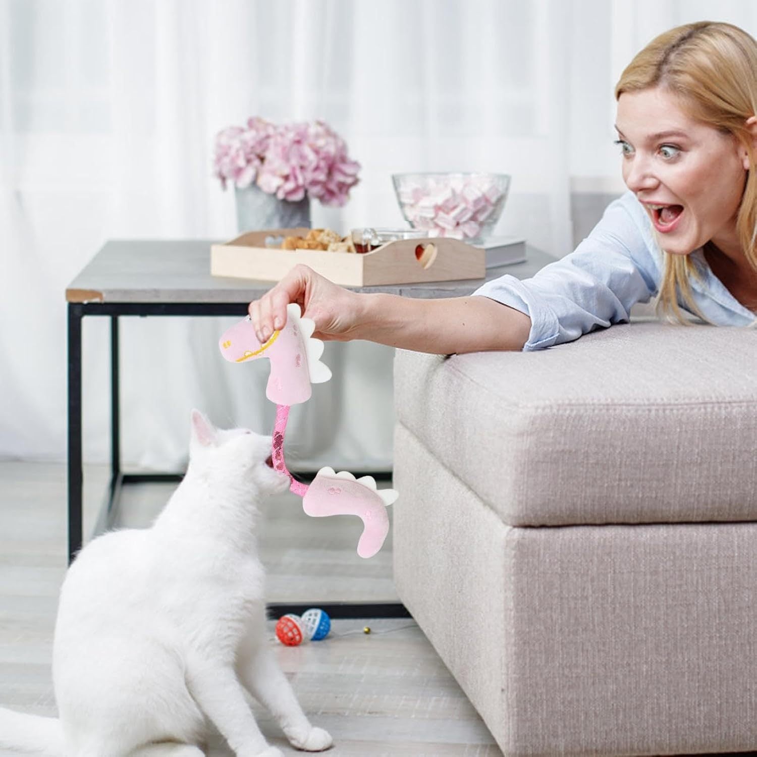 Person engaging a white cat with a pink plush dinosaur cat toy on a beige couch