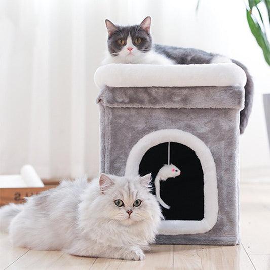 Two cats near a gray plush cat cave with white trim; one cat resting on top and the other lying in front of the entrance with a hanging mouse toy inside