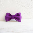 Load image into Gallery viewer, Purple velvet dog bow tie displayed on white wood surface with textured wall background.
