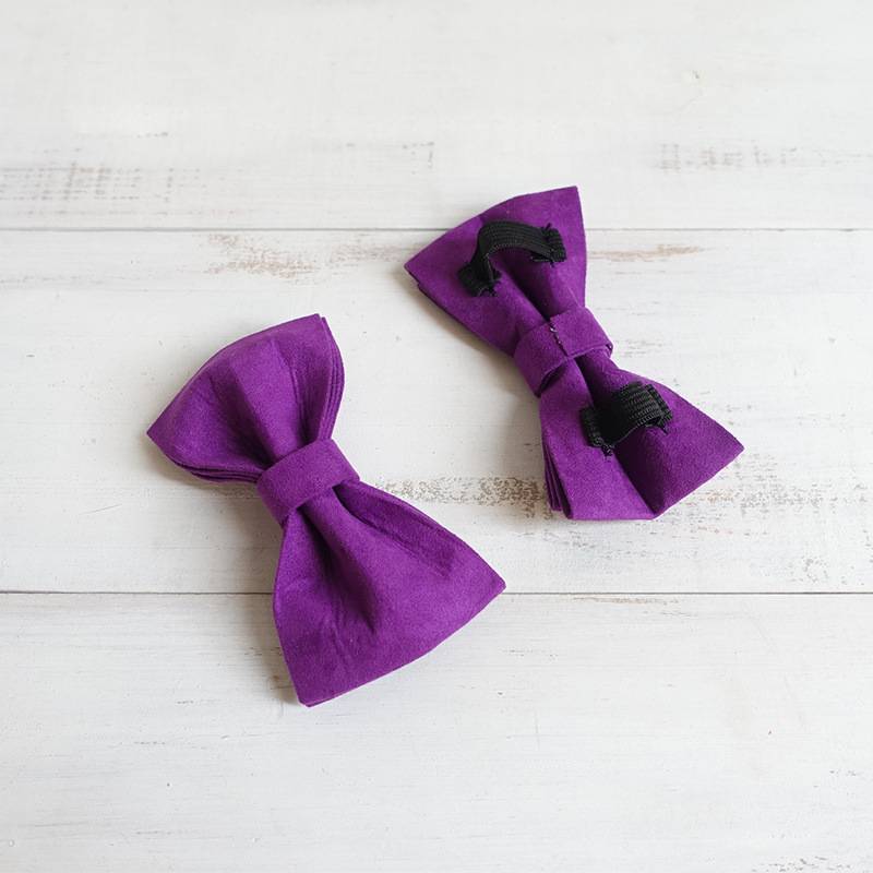 Two purple velvet dog bow ties showing back with black elastic loops on white wood surface.