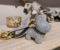 Charger l'image dans la visionneuse de la galerie, Close-up of a white crystal dog keychain with a gold keyring and crystal-embellished wrist strap on a light wood surface
