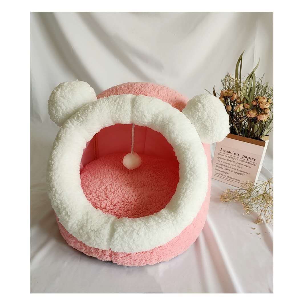 Pink and white plush cat cave bed with round ears and pompom toy on white surface next to dried flowers