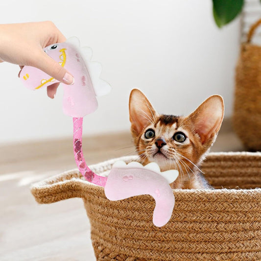 Small striped kitten watching a person hold up a pink plush dinosaur cat toy inside a wicker basket