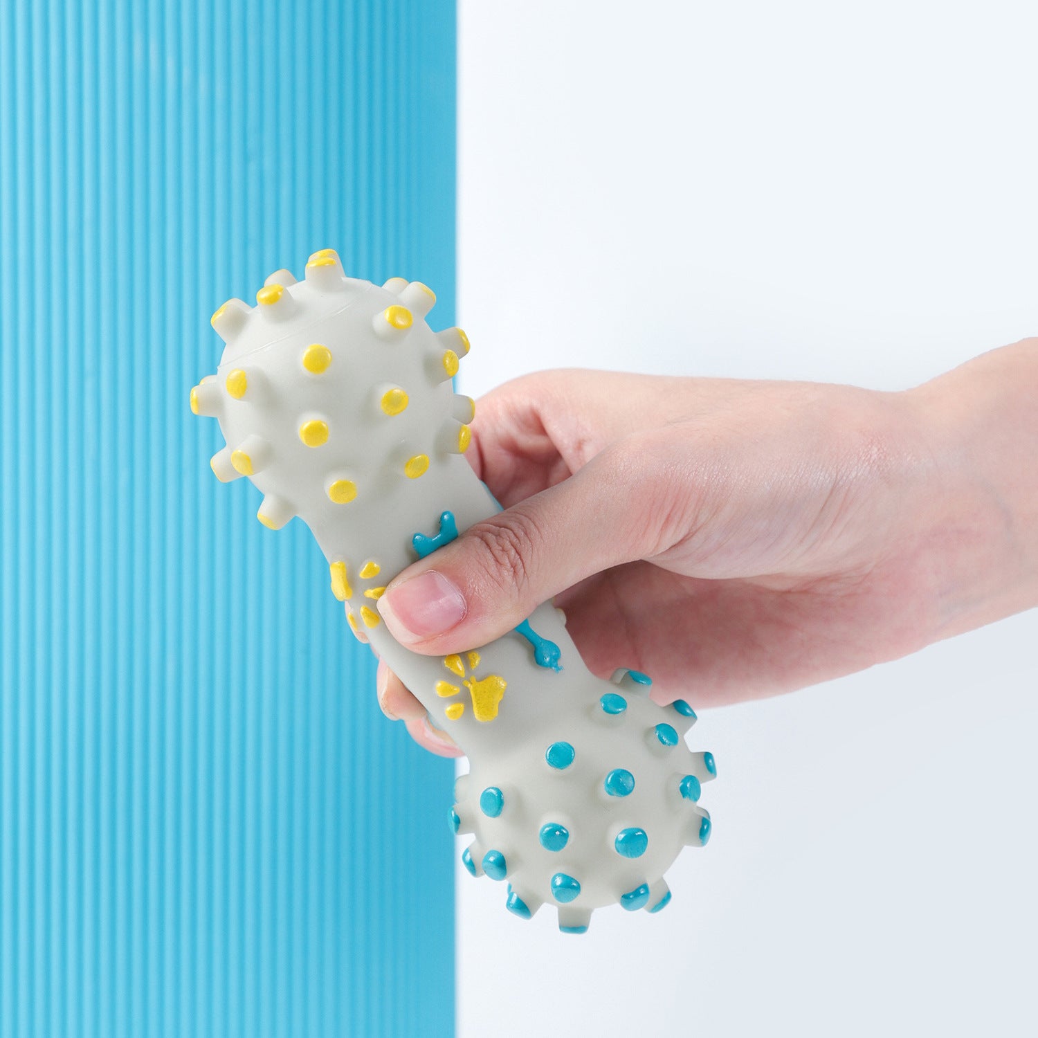 Hand holding a bone-shaped dog chew toy with yellow and blue massage nodes against white and blue background