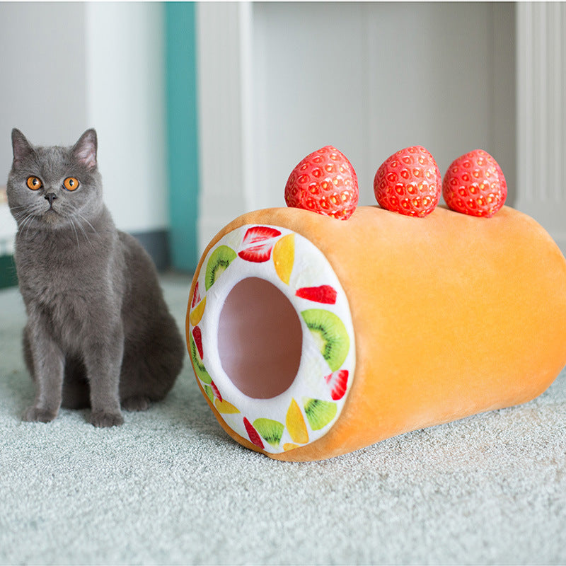Gray cat sitting beside the orange cake roll shaped plush cat tunnel with strawberry decorations