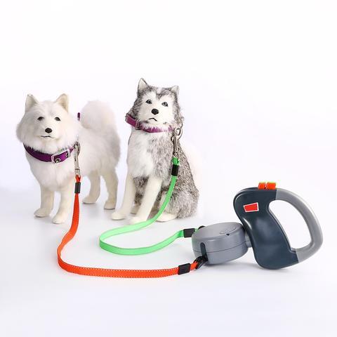 Dual retractable dog leash attached to two dogs with orange and green nylon straps and ergonomic handle on white background