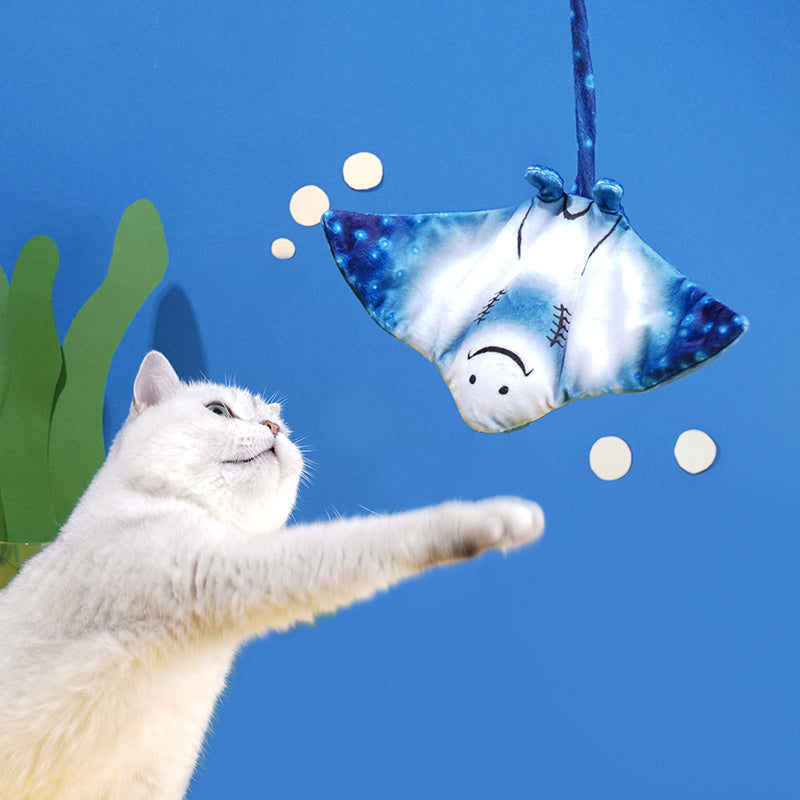White cat reaching out to touch a hanging blue manta ray plush cat toy against a blue background with green seaweed decorations