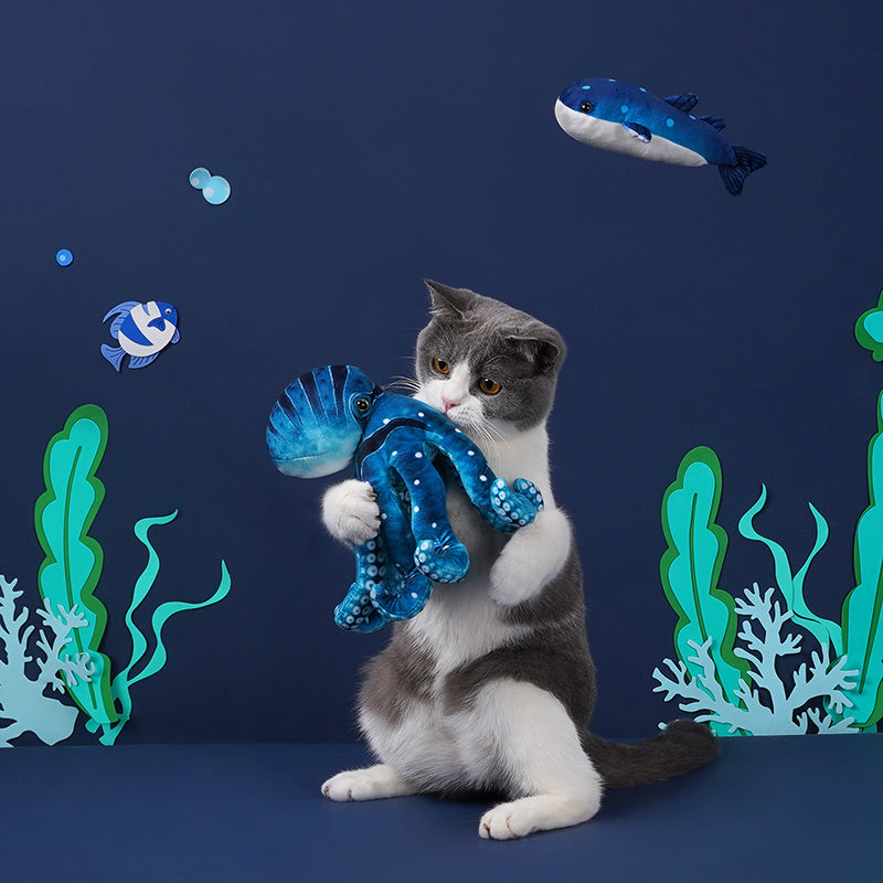 Gray and white cat holding a blue octopus plush cat toy in a playfully decorated underwater scene
