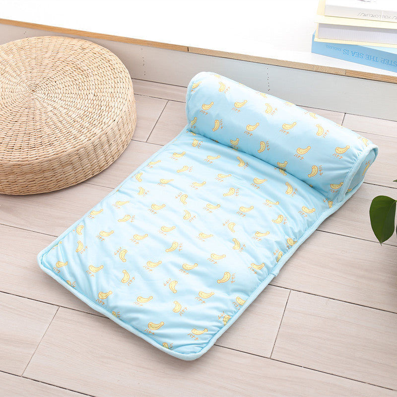 Light blue cooling pet mat with yellow banana pattern and attached bolster pillow displayed on wooden floor next to natural woven cushion