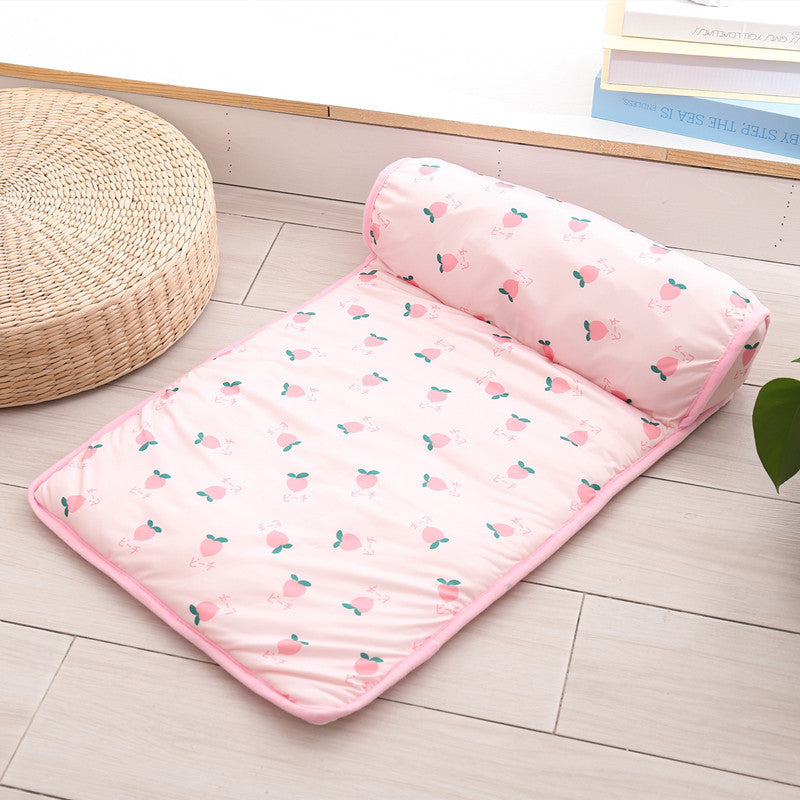 Pink cooling pet mat with peach fruit print and attached bolster pillow shown on wooden floor beside a woven cushion