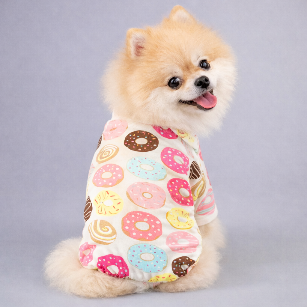 Small fluffy dog wearing white pajamas covered in various colorful donuts pattern, sitting and looking back happily