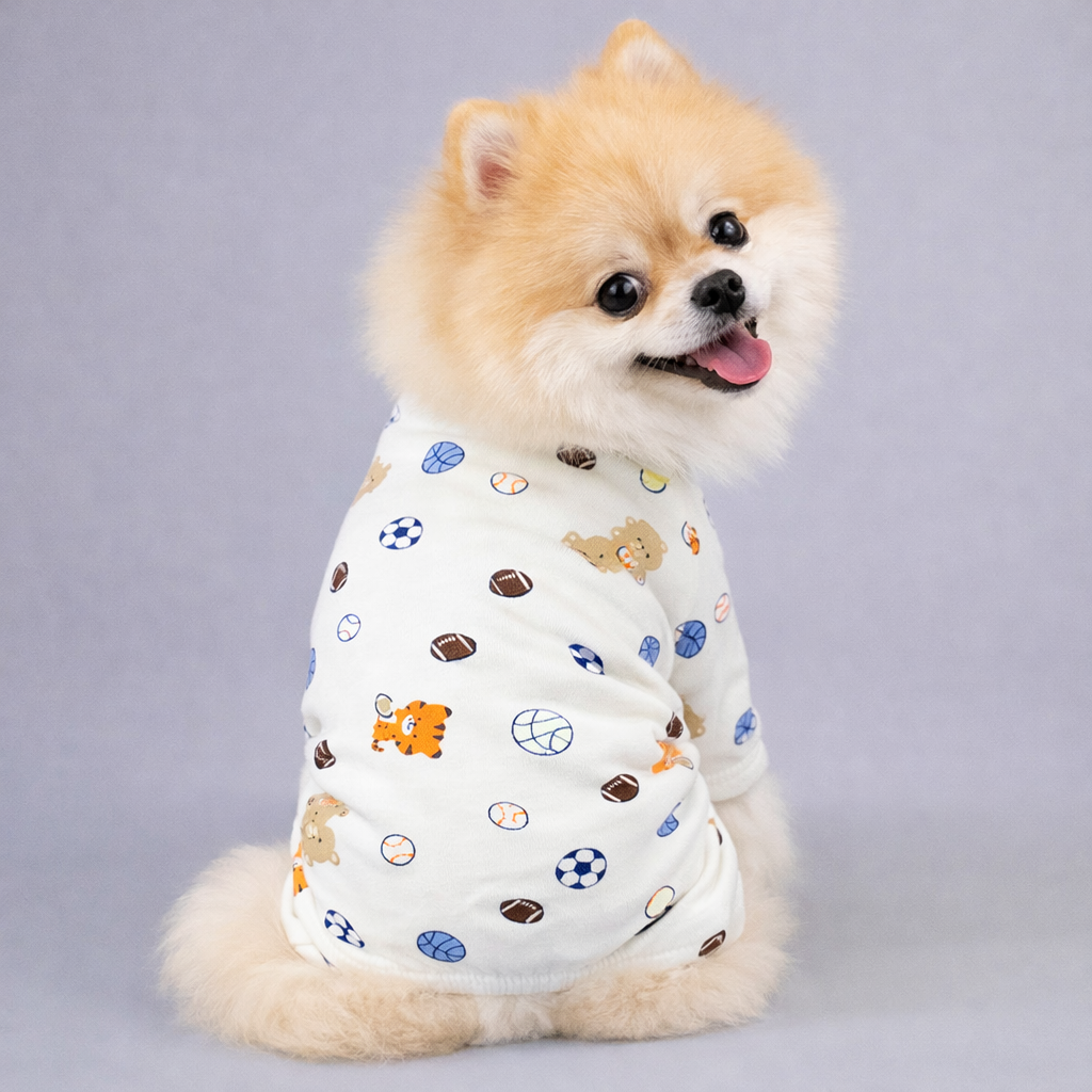 Small fluffy dog wearing white pajamas with sports balls and animal prints, sitting and looking at camera