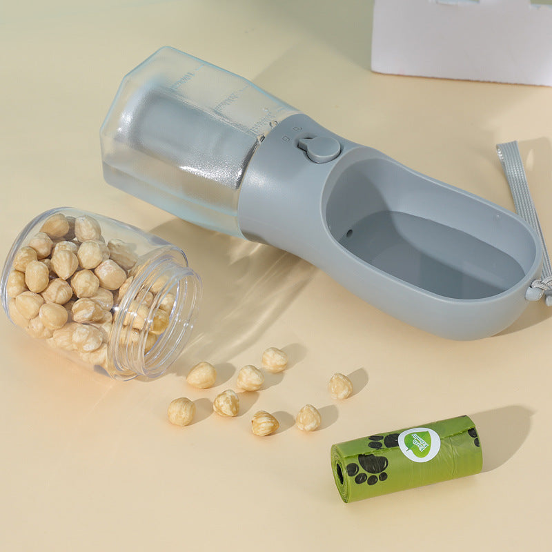 Portable dog feeder bottle lying flat with detachable food container spilling dry kibble and green dog waste bag roll nearby