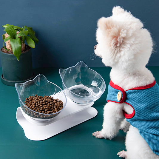 White elevated pet feeder with two clear cat ear shaped bowls, one filled with pet kibble, beside a small white dog in blue harness