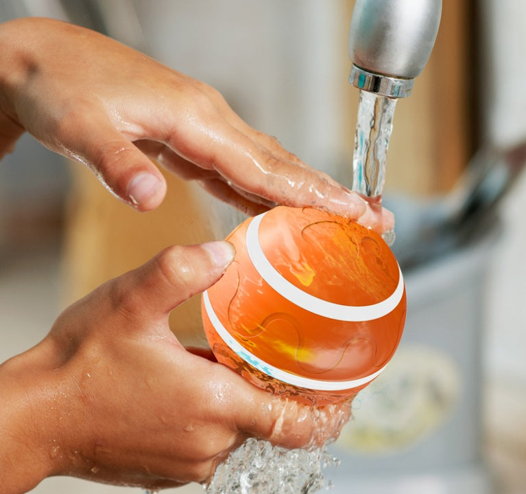 Hands washing an orange intelligent cat ball under running water faucet
