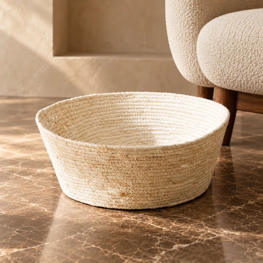 Open woven cat bed in a natural cream colour on brown marble beside a cream upholstered chair.