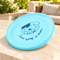 Cargar la imagen en la vista de la galería, Light blue dog flying disc with the original dog-and-bone print on a light ivory-beige marble terrace table.
