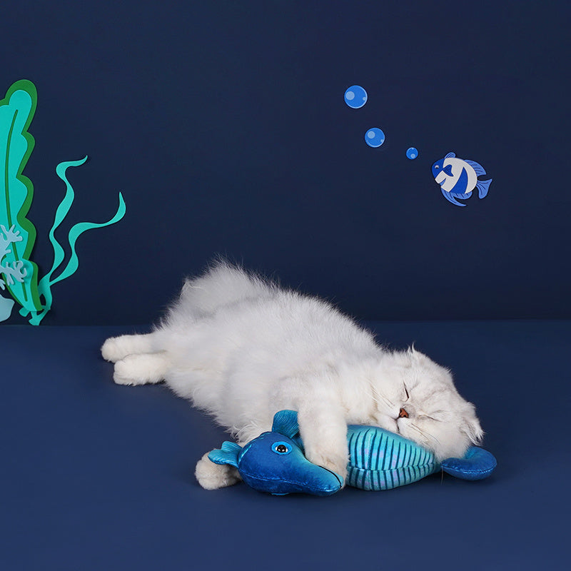 White fluffy cat cuddling with a blue seahorse plush toy on a dark blue surface with ocean-themed paper cutouts