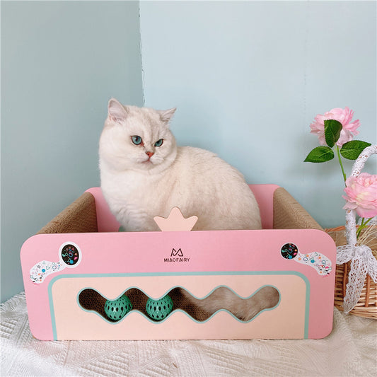 White cat sitting inside pink crown-shaped corrugated cat scratcher bed next to flowers